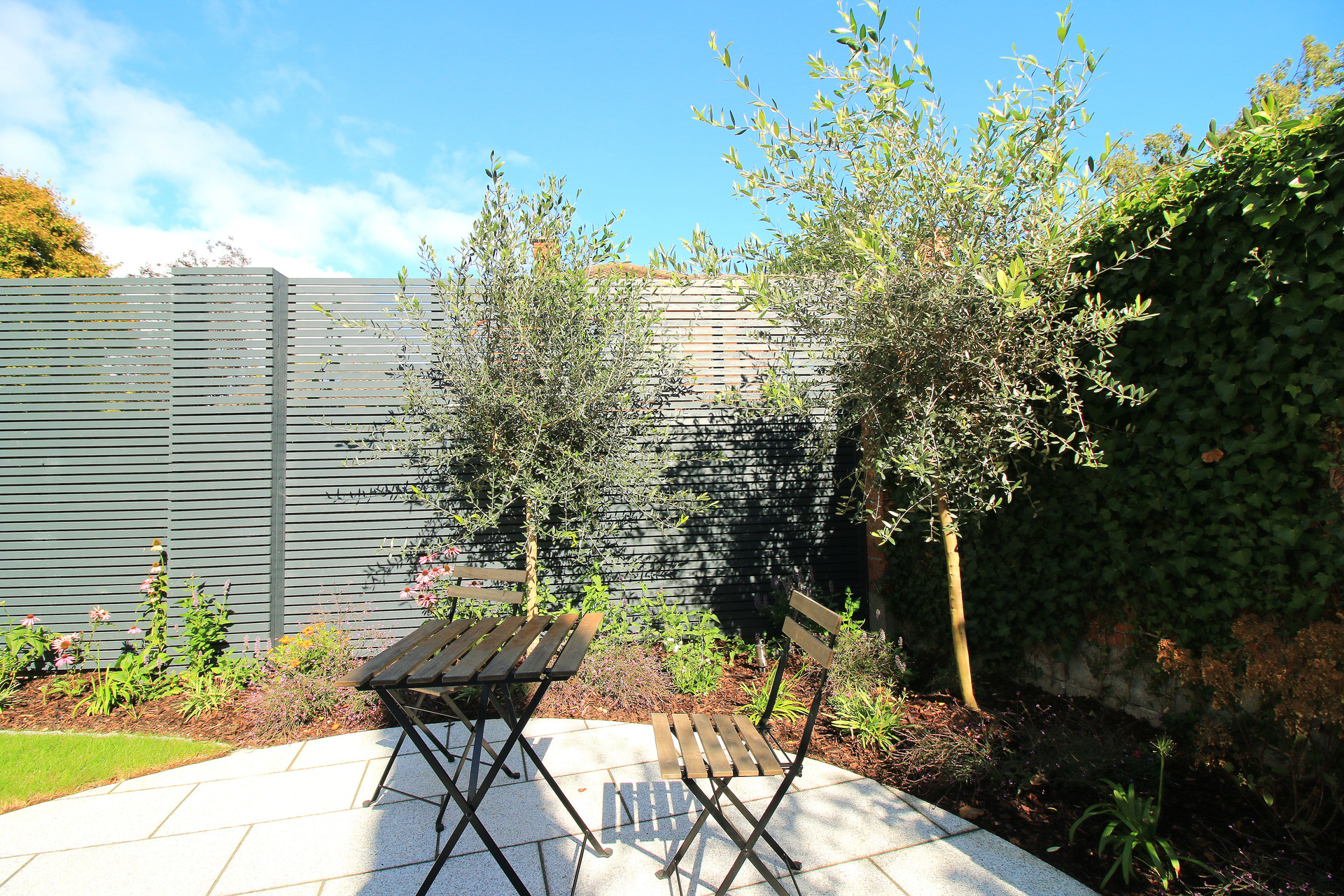 Family Garden Design & Landscaping in Blackrock, Co Dublin includes custom made horizontal slatted fencing, granite patio and paving, granite sett edged planted borders & mowing edges, stainless steel garden spike lighting, Classic Garden Elements Bagatelle Rose Arch, and a stunning planting scheme of diverse specimen olive trees, mature shrubs & perennials.
